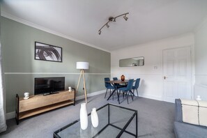 Un salon moderne doté d’un téléviseur à écran plat, d’une console en bois, d’une table basse en verre et d’un coin repas avec des chaises bleues et une table en bois.