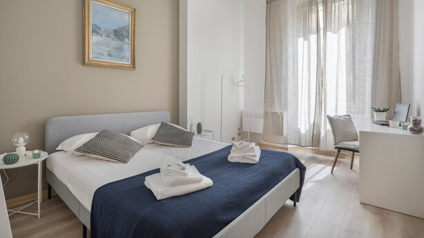 A modern hotel room with a large bed, a desk, and a chair. There is a painting on the wall, a window with curtains, and a small table with a lamp and a plant. - Synagogue Suite (Florence)