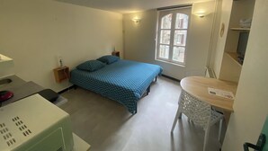 Un dormitorio pequeño y luminoso con una cama individual, un escritorio y una silla. Se ve una ventana con vistas a edificios.