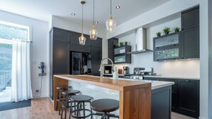 Une cuisine moderne dotée d’un îlot central, de tabourets de bar, de meubles de rangement aux couleurs sombres et d’un éclairage suspendu.