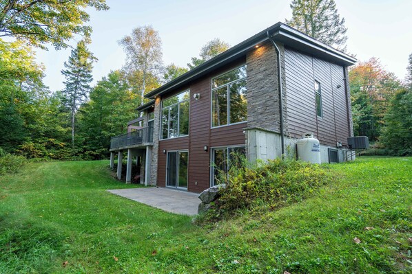 A modern two-story house with a large deck, surrounded by lush greenery and tall trees.