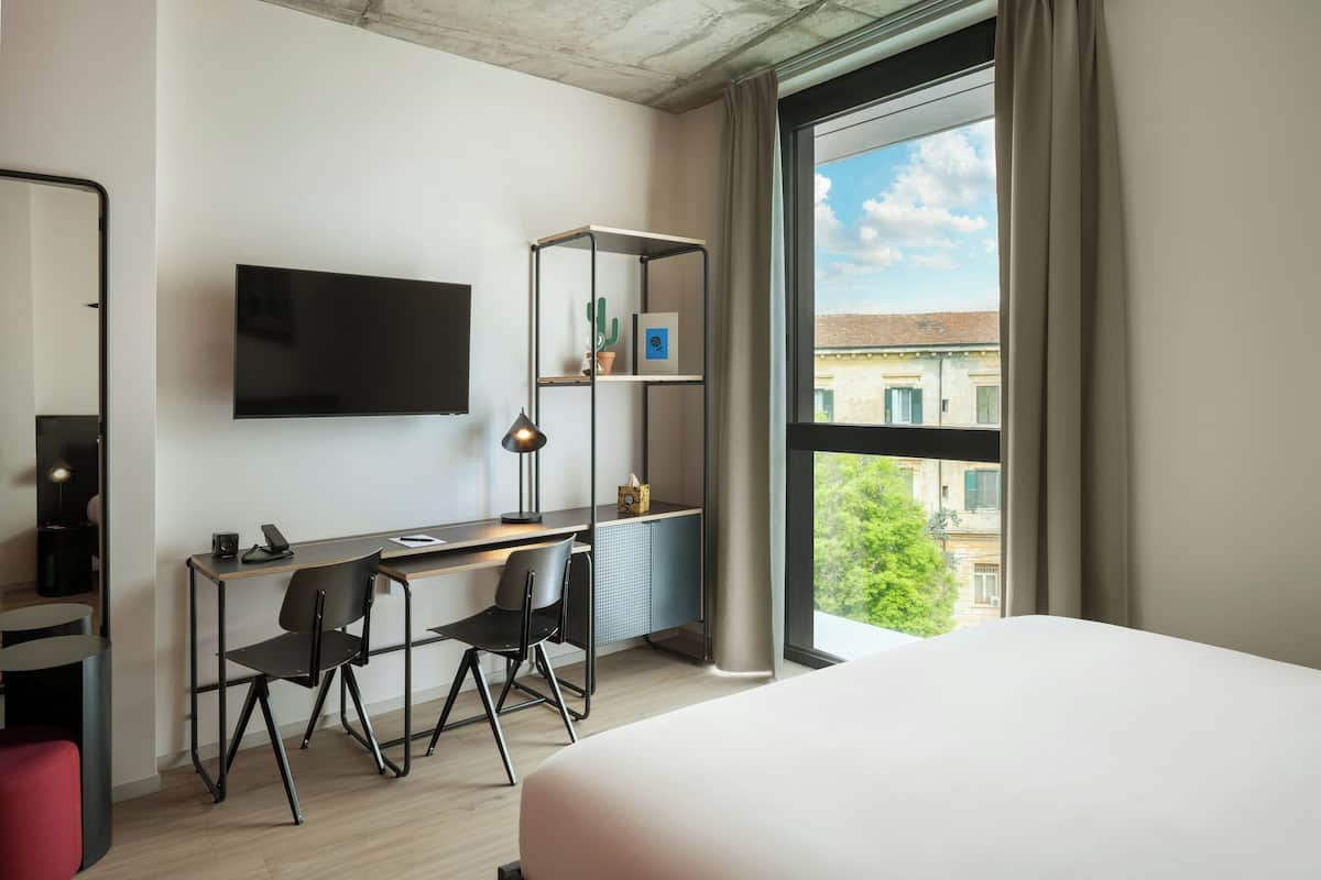 A modern hotel room with a bed, desk, chairs, a TV, and a large window with a view of buildings.