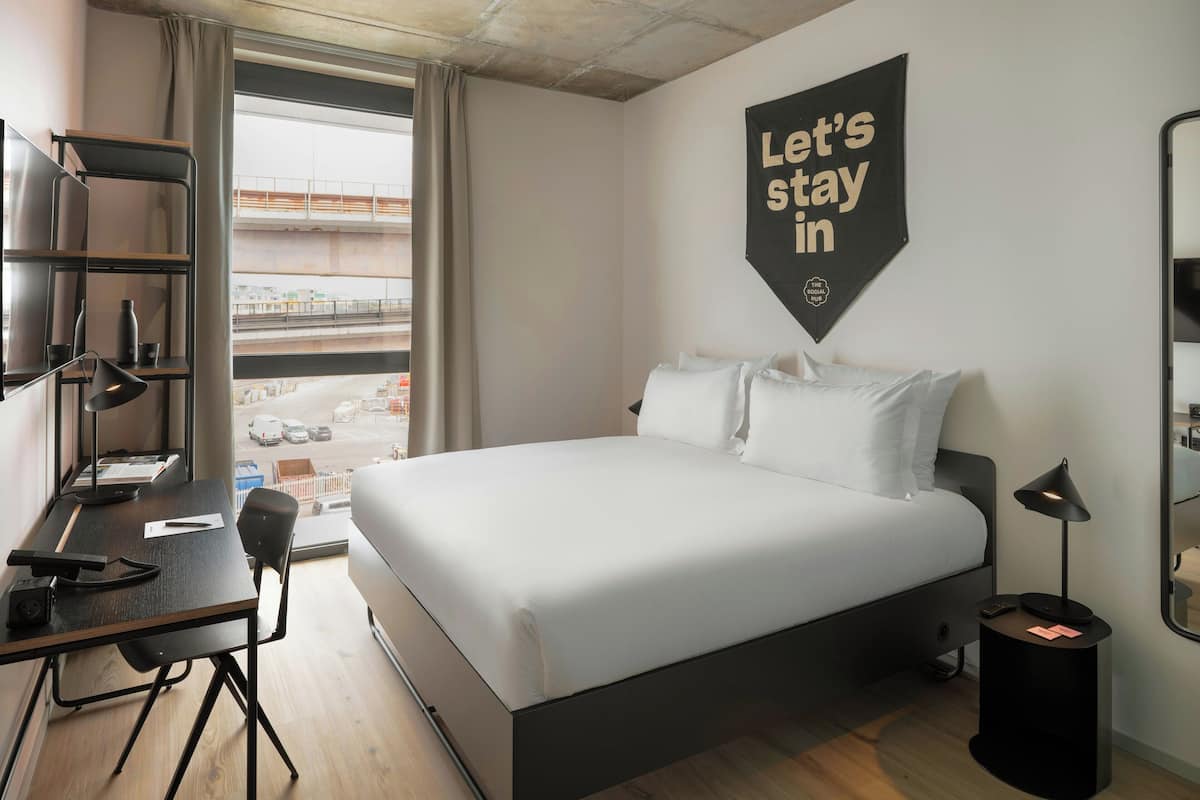 A hotel room with a bed, desk, chair, and a view of the city through the window.
