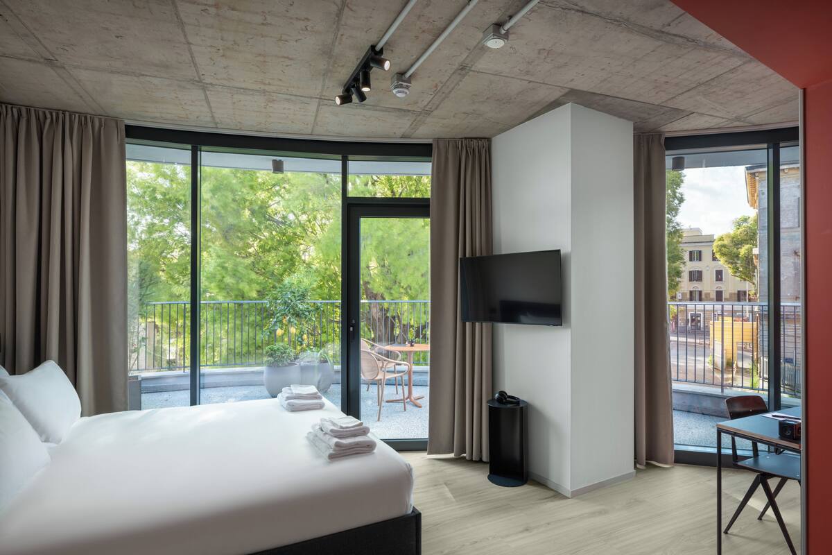A modern hotel room with a large bed, a flat-screen TV, a balcony with a view of the city, and a small table with a chair.