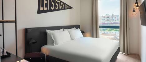 Ein Hotelzimmer mit einem großen Bett, einem Schreibtisch mit Telefon und Kamera, einem Schild mit der Aufschrift "LET'S STAY IN" an der Wand und Blick auf Gebäude durch das Fenster.