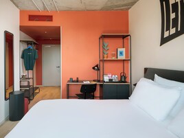 A modern hotel room with a large bed, a desk with a chair, a small shelf with decorative items, and a wardrobe.