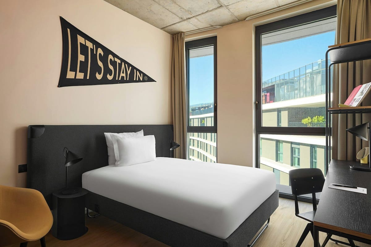 A hotel room with a bed, a desk, a chair, a lamp, and a large window with a view of buildings.