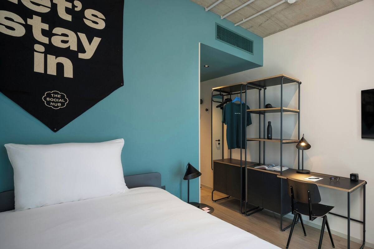 A modern hotel room with a bed, a desk with a chair, a hanging sign, and a wardrobe.
