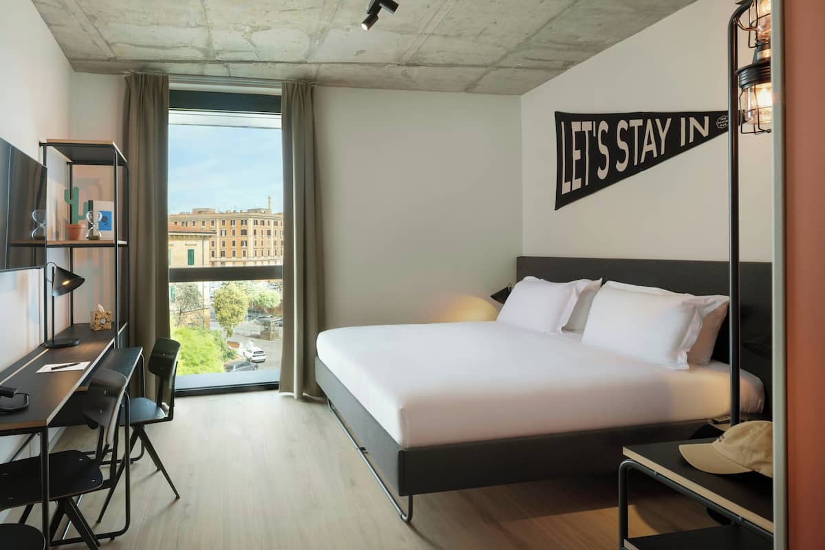 A hotel room with a bed, desk, chair, and a view of buildings outside.