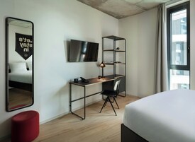 Et moderne hotelværelse med en seng, et skrivebord med stol, et fjernsyn og en rød puf.
