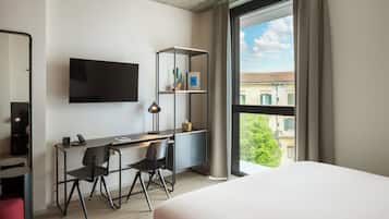 Una habitación de hotel moderna con una cama grande, un escritorio con una silla, un televisor montado en la pared y una ventana con vistas a edificios y árboles.