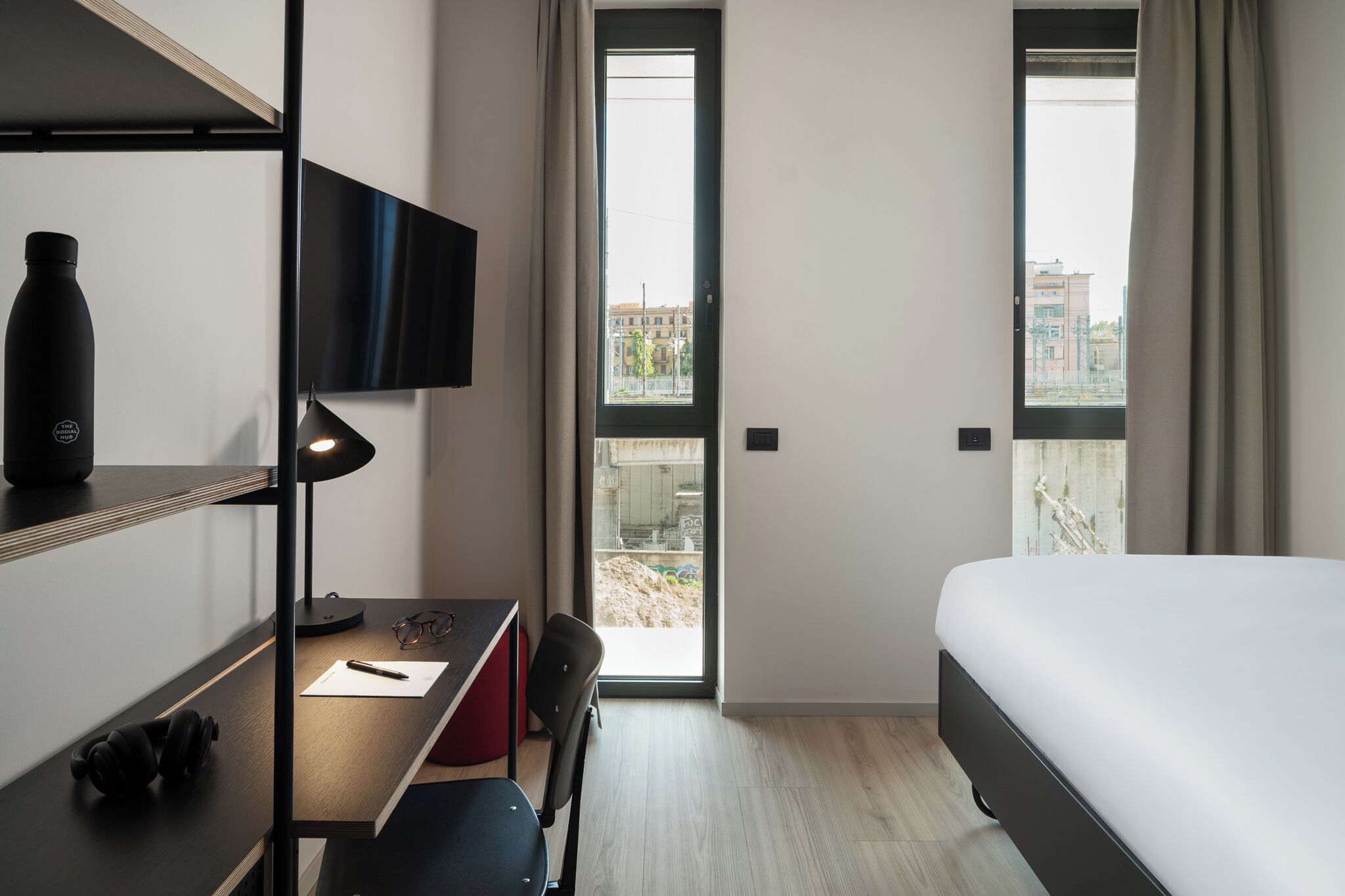 A hotel room with a bed, desk, chair, TV, and large windows offering a view of buildings outside.