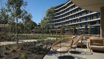 Um edifício moderno com fachada curva, área de estar externa com cadeiras de vime e um jardim arborizado.
