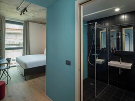 Et hotelværelse med seng, skrivebord, stol, bruseområde med glasdør og udsigt ud.