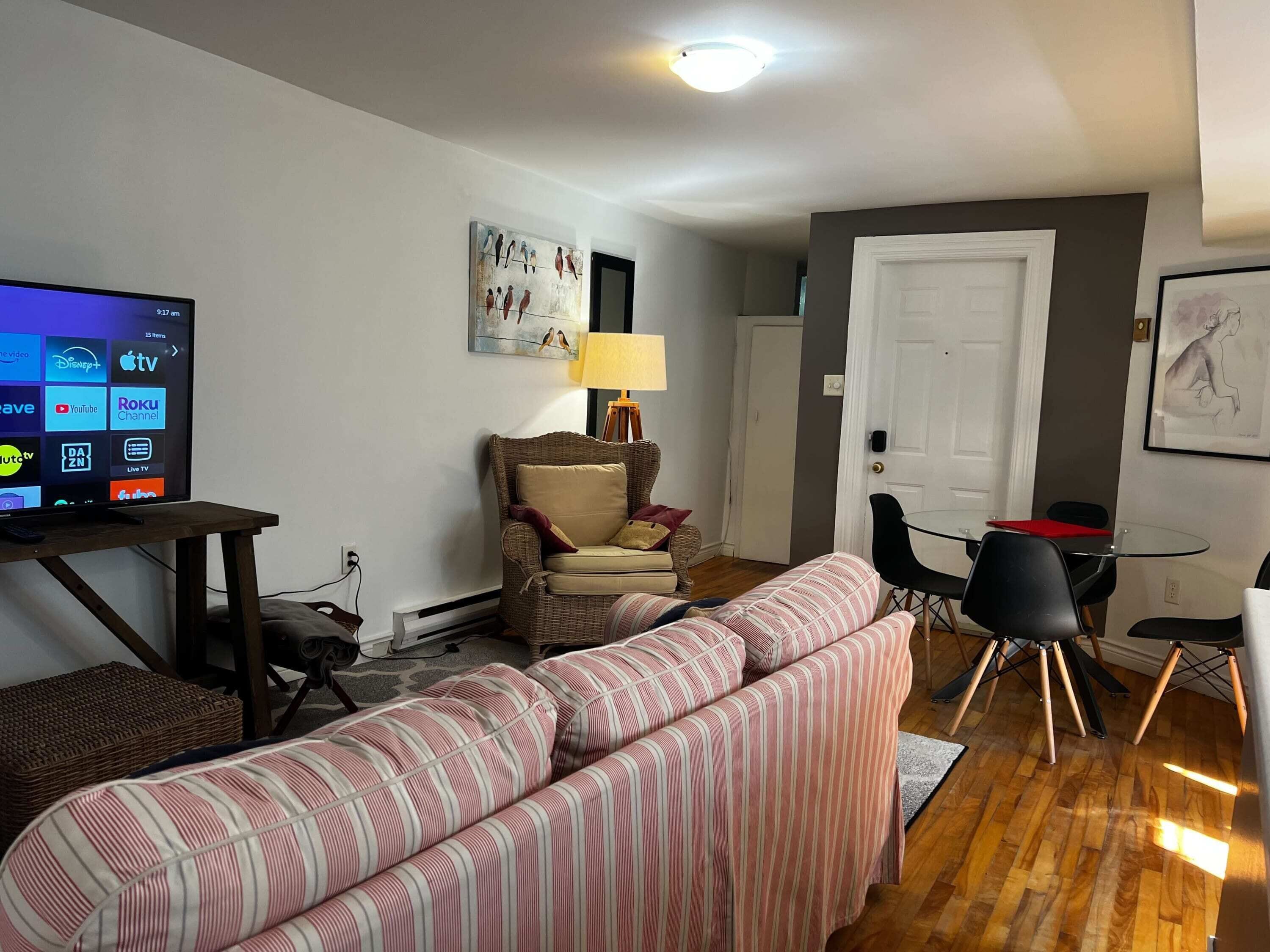 Un salon avec un téléviseur diffusant des services de streaming, un fauteuil en osier, un canapé à rayures, une table à manger avec des chaises et une porte.