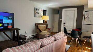 Un salon avec un téléviseur diffusant des services de streaming, un fauteuil en osier, un canapé à rayures, une table à manger avec des chaises et une porte.