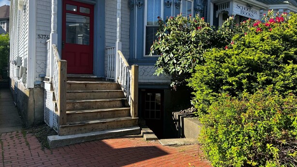 Un edificio de dos plantas con una puerta principal roja, un camino de ladrillo que conduce a la entrada y un pequeño jardín con arbustos verdes y flores.
