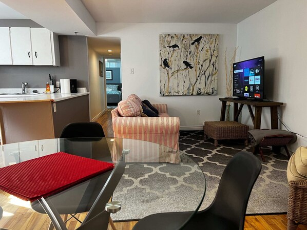 A modern living room with a glass dining table, a sofa, a TV, and a painting on the wall.