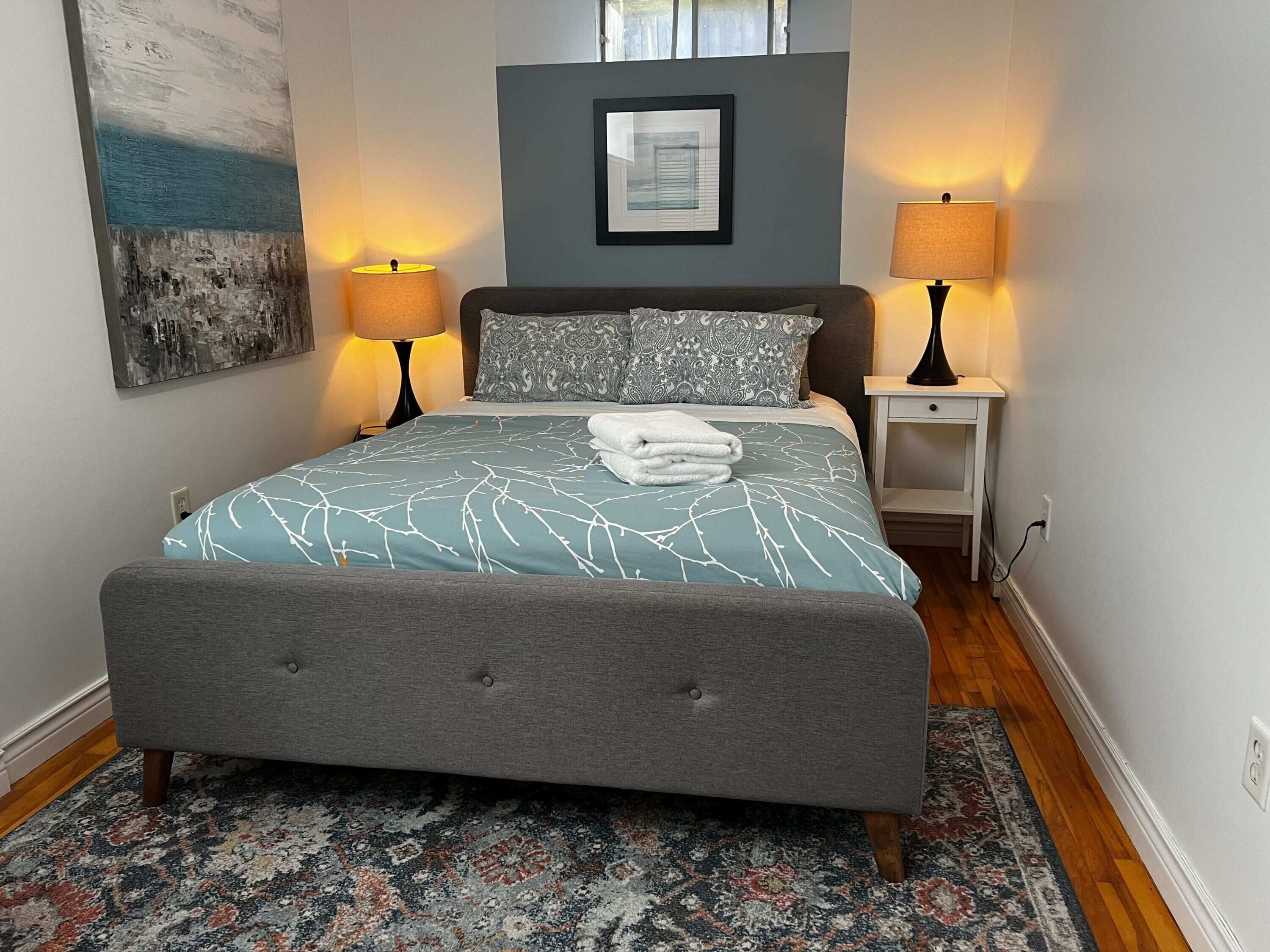 Un lit bien fait, avec une tête de lit rembourrée grise, une couvre-lit bleue et blanche à motifs, et une serviette blanche pliée posée sur le lit. De chaque côté du lit, on trouve deux lampes de chevet, une petite table de chevet blanche et un tableau encadré au-dessus du lit.
