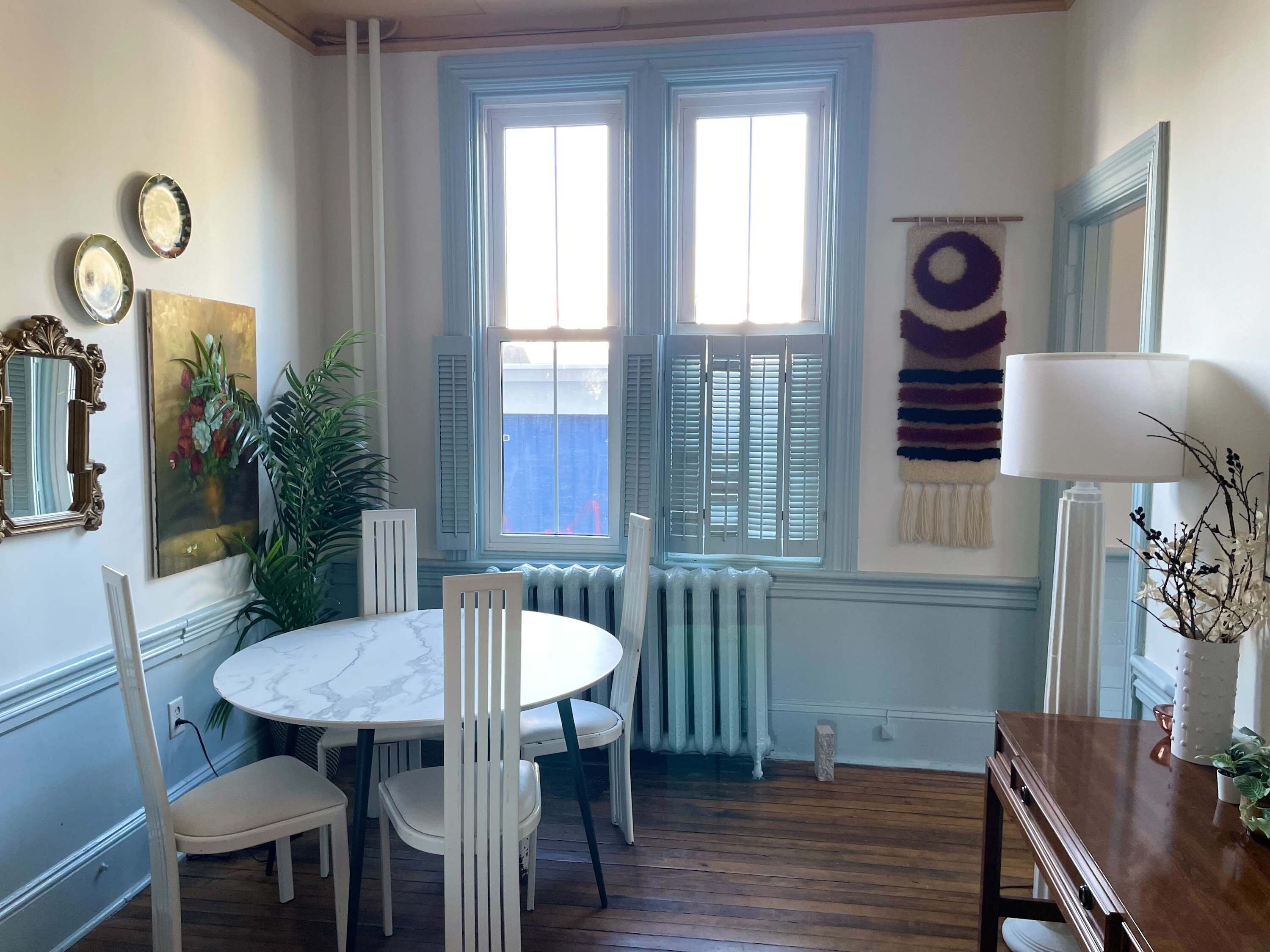A dining area with a round table and white chairs, a radiator, and a window with shutters.