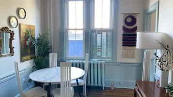 A dining area with a round table and white chairs, a radiator, and a window with shutters.