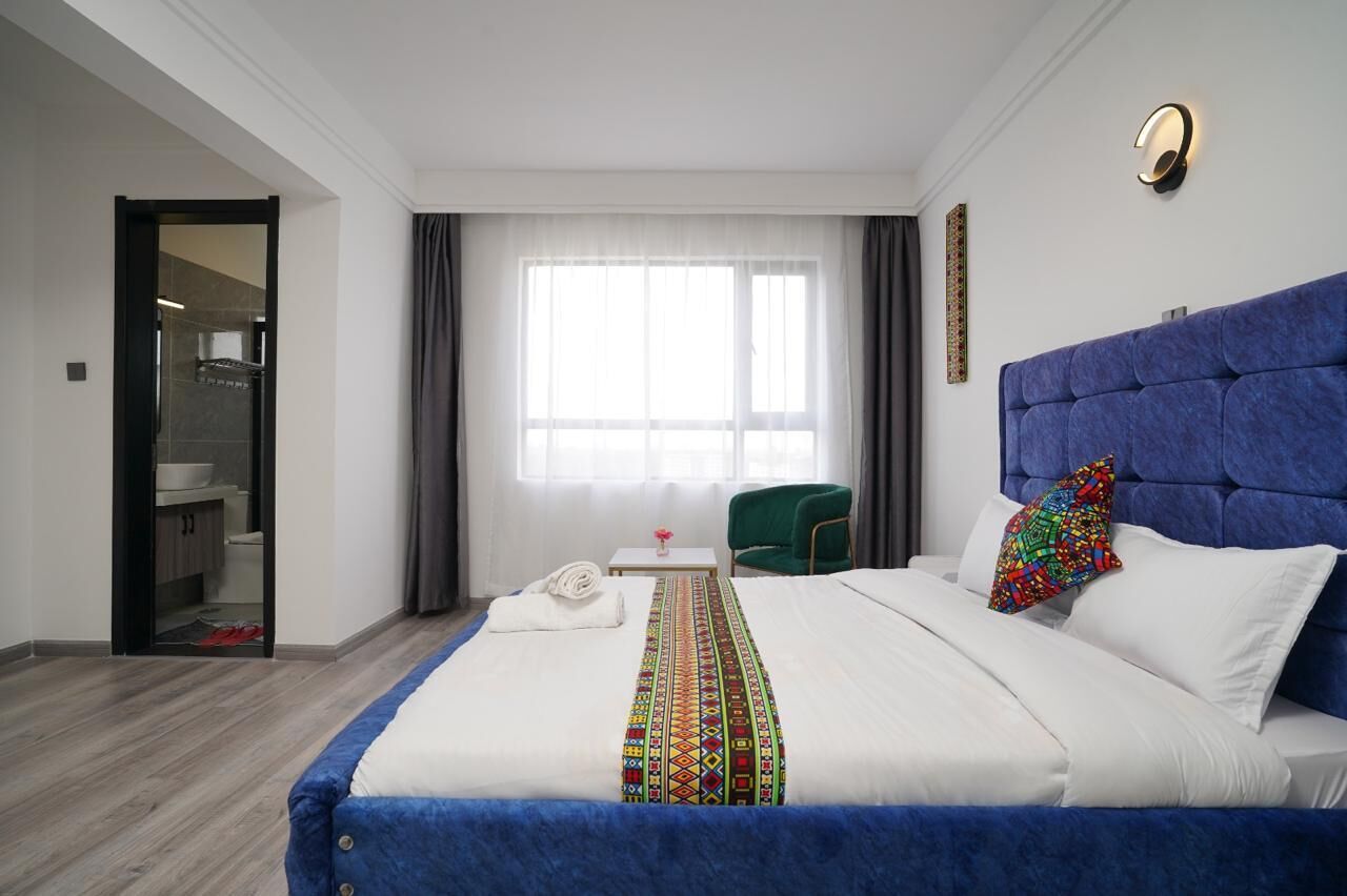 Ein Hotelzimmer mit einem großen Bett, einem grünen Sessel, einem kleinen Tisch mit einer Vase und einem durch eine offene Tür einsehbaren Badezimmer.