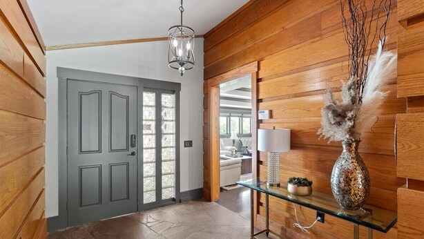 Un pasillo revestido de madera con una puerta gris, una mesa consola de cristal con un jarrón decorativo y una lámpara colgante.