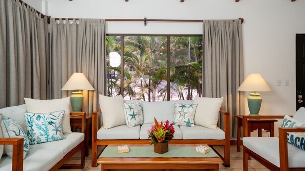 A living room with a sofa, two armchairs, a coffee table, and two lamps. There is a window with curtains and a view of palm trees. - Sunny Days Villa At Playa Hermosa (Playa Hermosa)