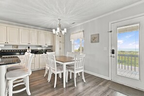 Une cuisine avec des meubles blancs, une table à manger avec des chaises, un lustre et une vue sur l’extérieur, visible à travers une porte.