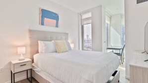 A modern hotel room with a large bed, a nightstand with a lamp, a city view through the window, and a painting on the wall.
