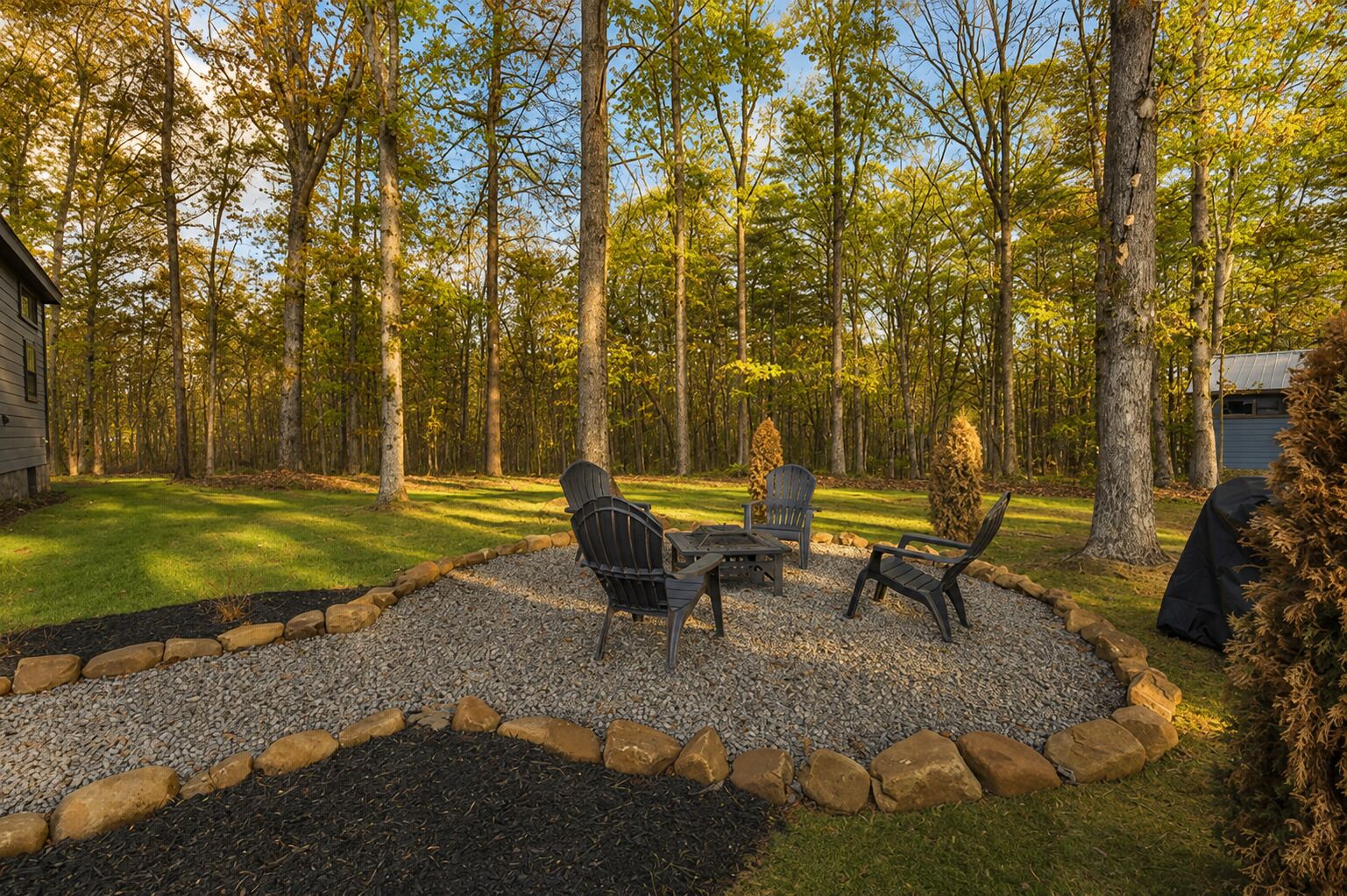 Une terrasse aménagée avec du mobilier d’extérieur, entourée d’une zone recouverte de gravier et bordée de pierres, située dans un espace boisé.