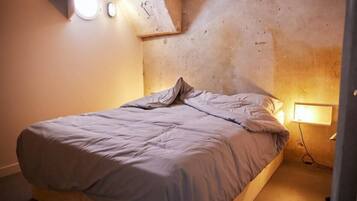 Ein kleines Zimmer mit einem Einzelbett, einer Betonwand, einer Nachttischlampe und einer Steckdose.