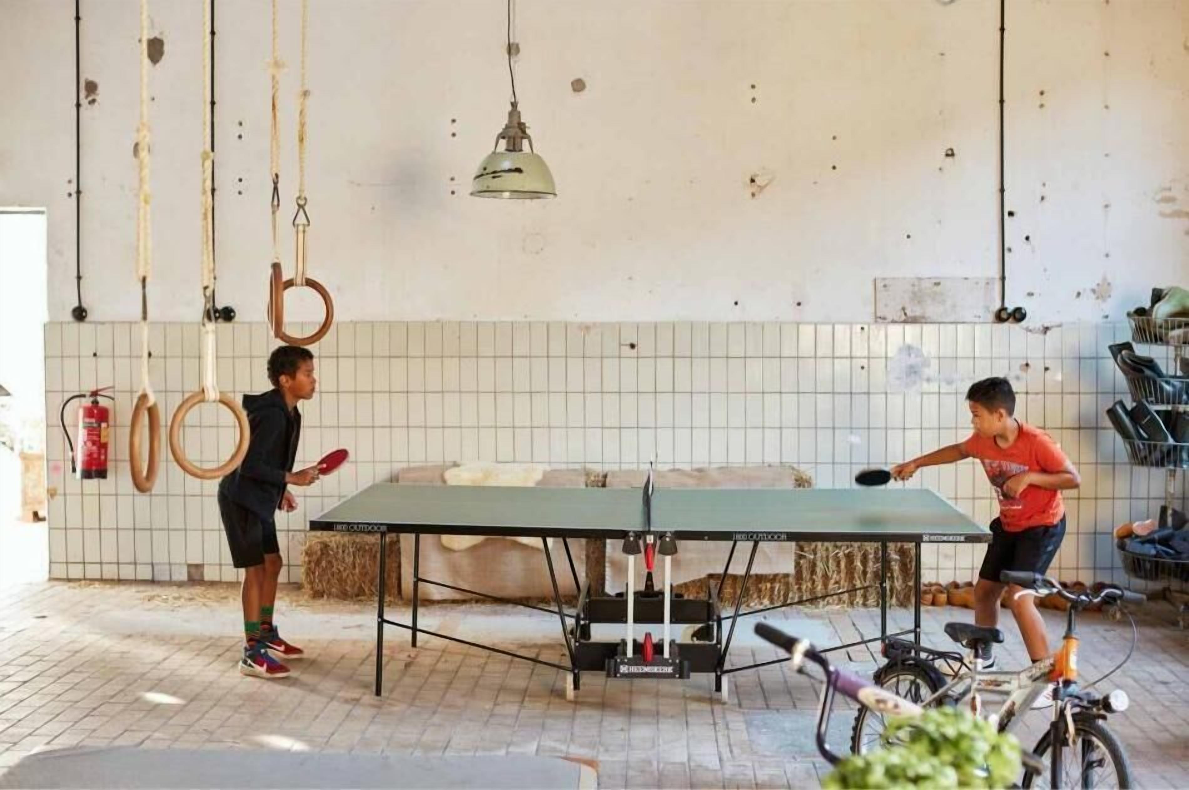 Two people playing table tennis in a room with a ping pong table, exercise equipment, and a bicycle.