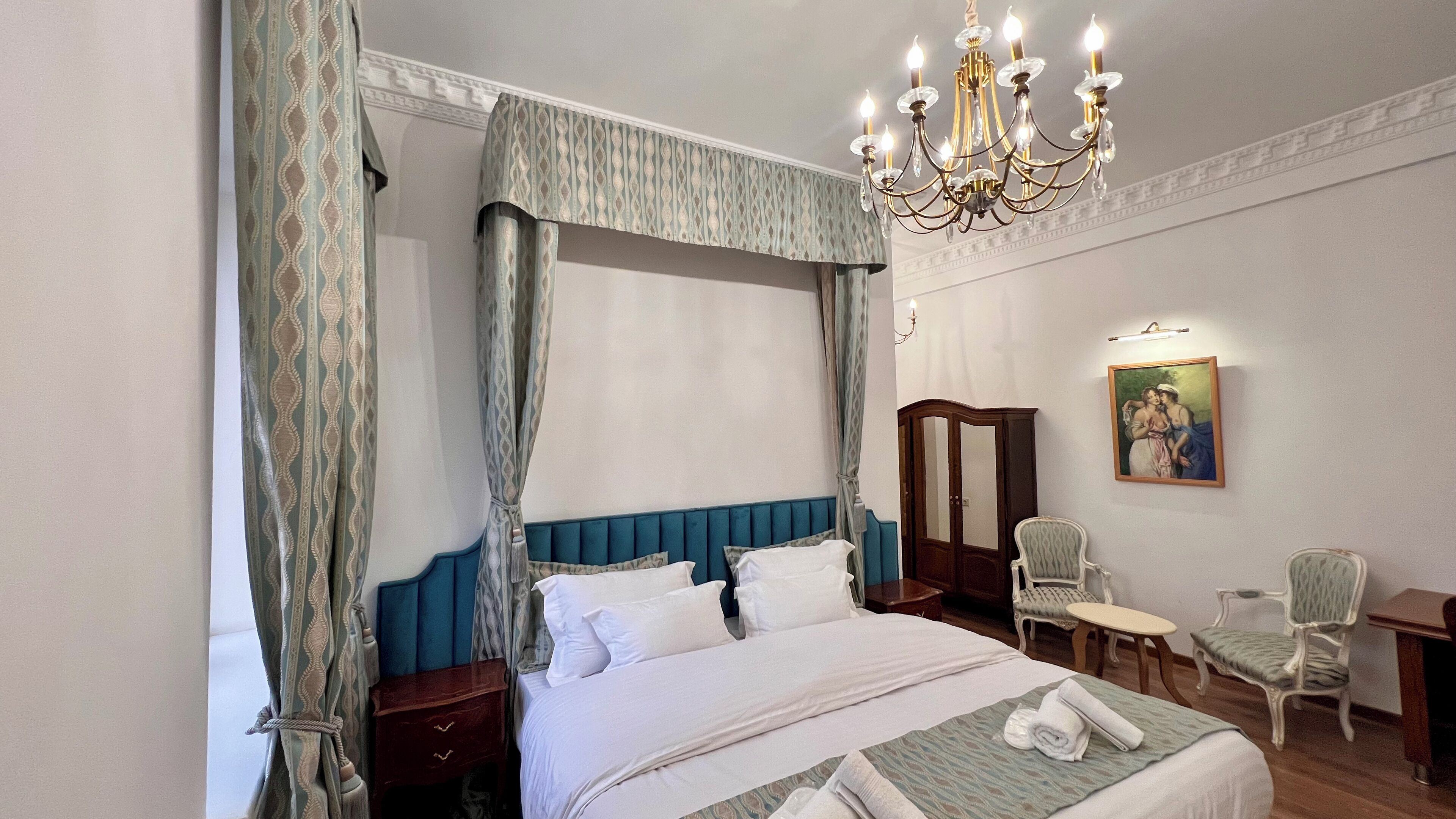 A bedroom with a four-poster bed, a chandelier, a wardrobe, a chair, and a painting on the wall.