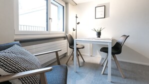 Una habitación pequeña con ventana, una mesa, dos sillas, una planta en maceta y un cuadro en la pared.