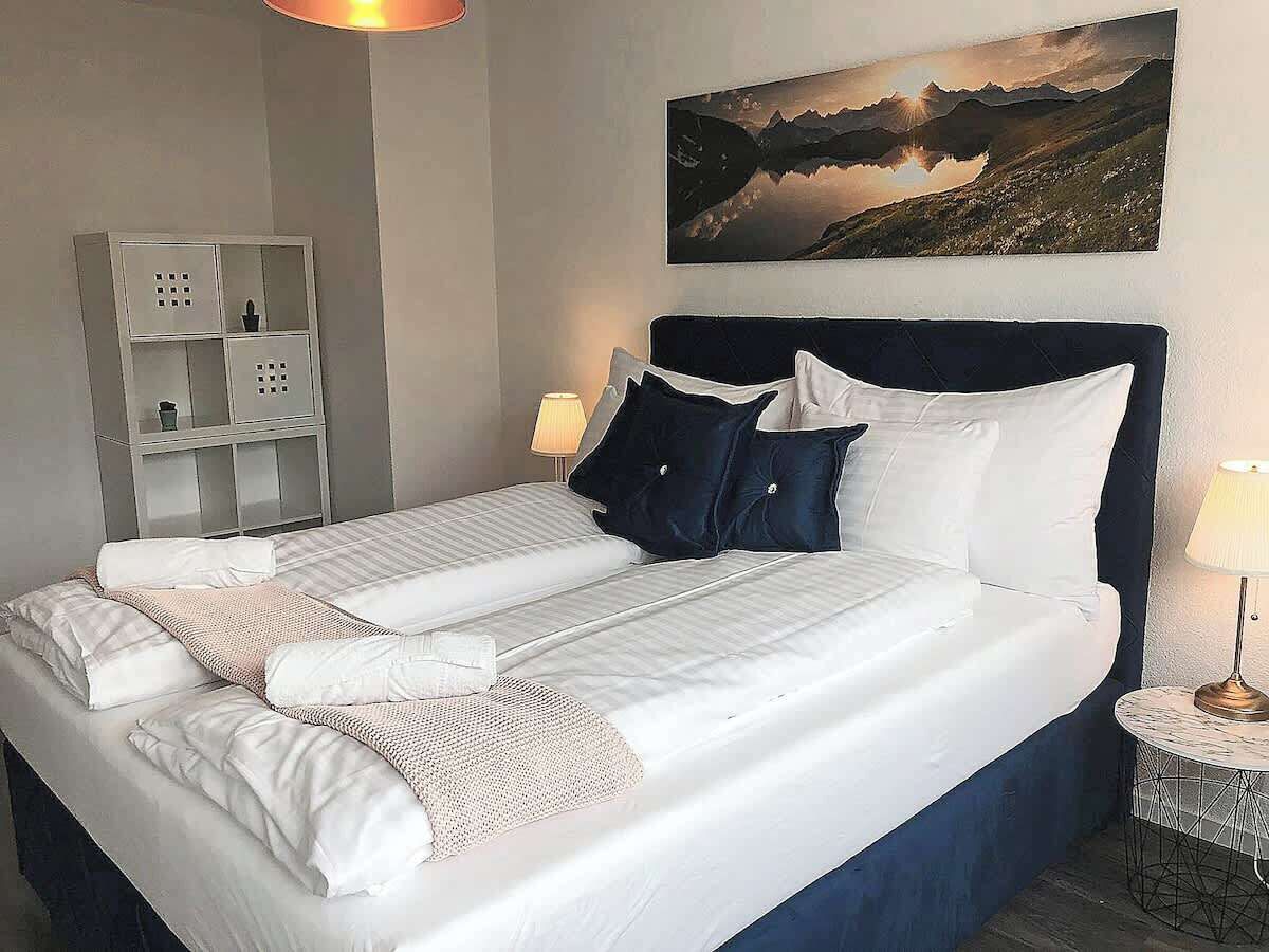 A double bed with white linens, a dark headboard, a painting of a mountain landscape, and a small bedside table with a lamp.