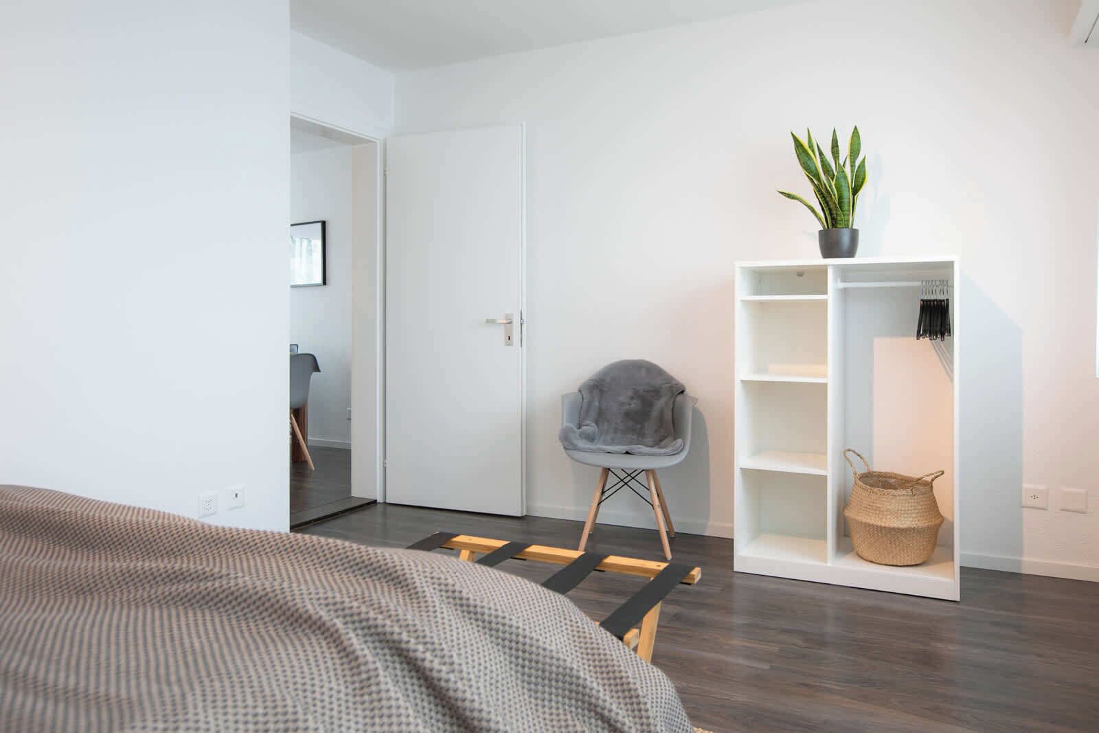 Ein modernes Zimmer mit einem Bett, einem grauen Sessel, einer weißen Regaleinheit mit einer Pflanze und einem gewebten Korb.