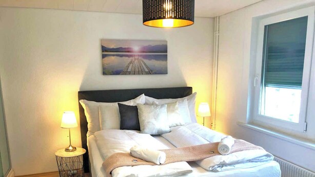 A neatly made bed with pillows and a blanket, two bedside lamps, a painting on the wall, and a window with blinds.