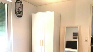 A room with a white wardrobe, a bed with a black headboard, and a small window with blinds.