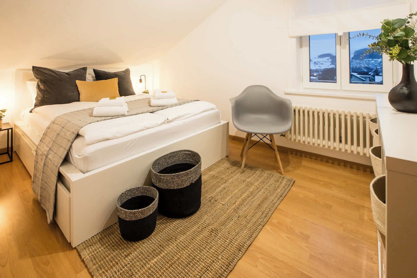 A bedroom with a bed, bedside tables, a chair, a radiator, and a window with a view.
