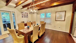 A dining area with a wooden table and wicker chairs, a chandelier, and a view of the outdoors through large windows. - Just minutes to the beach with a hot tub and fire pit! (Rehoboth Beach)