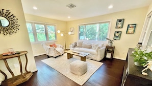 A living room with a white sofa, a patterned armchair, a coffee table, and a rug. There are two windows, a floor lamp, and wall art. - Just minutes to the beach with a hot tub and fire pit! (Rehoboth Beach)