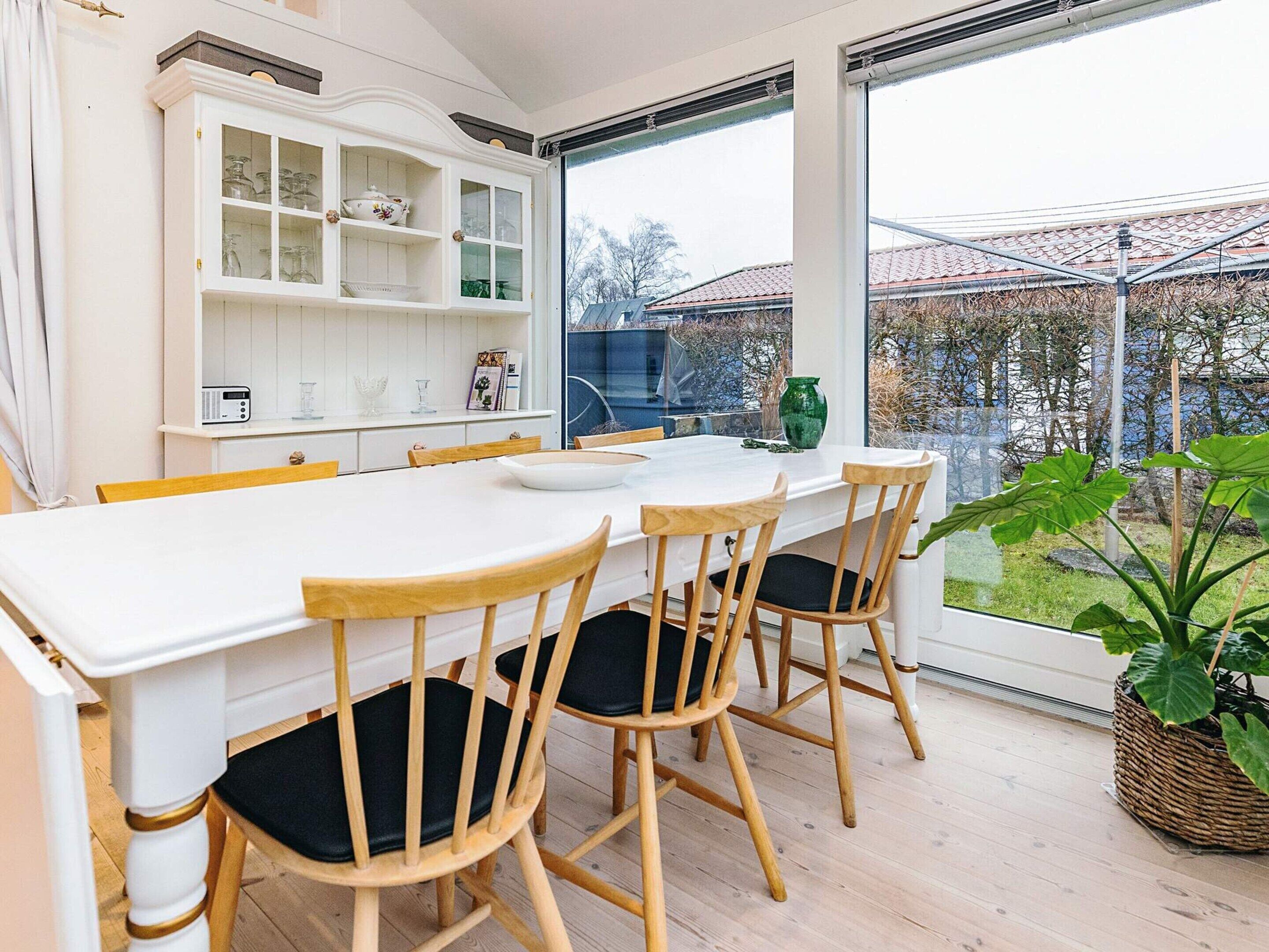 A modern kitchen with a white table, wooden chairs, and a large window overlooking a garden.