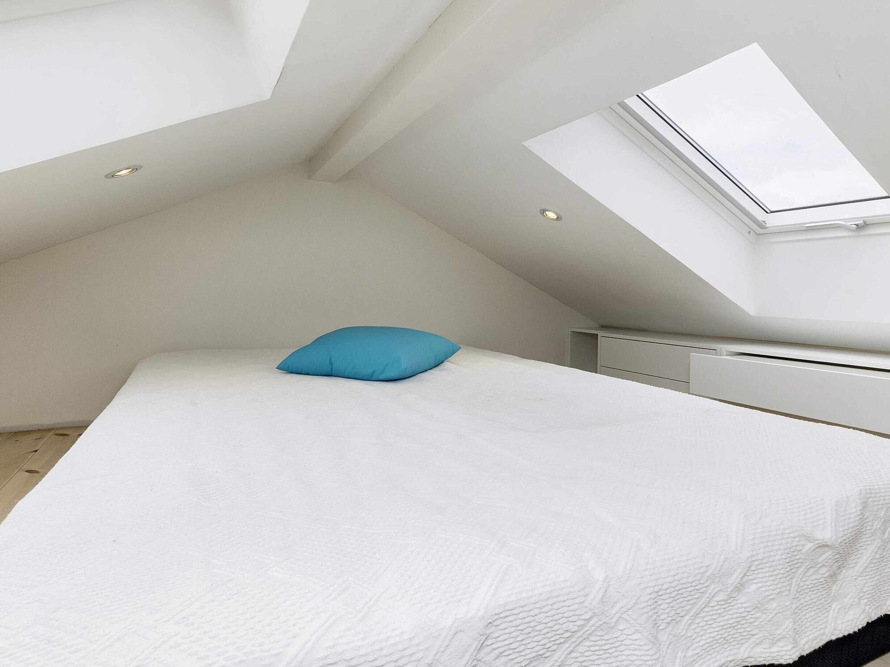 A bedroom with a sloped ceiling, a skylight, a bed with a white bedspread and a blue pillow, and a white radiator.