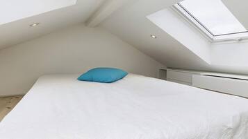 A bedroom with a sloped ceiling, a skylight, a bed with a white bedspread and a blue pillow, and a white radiator.