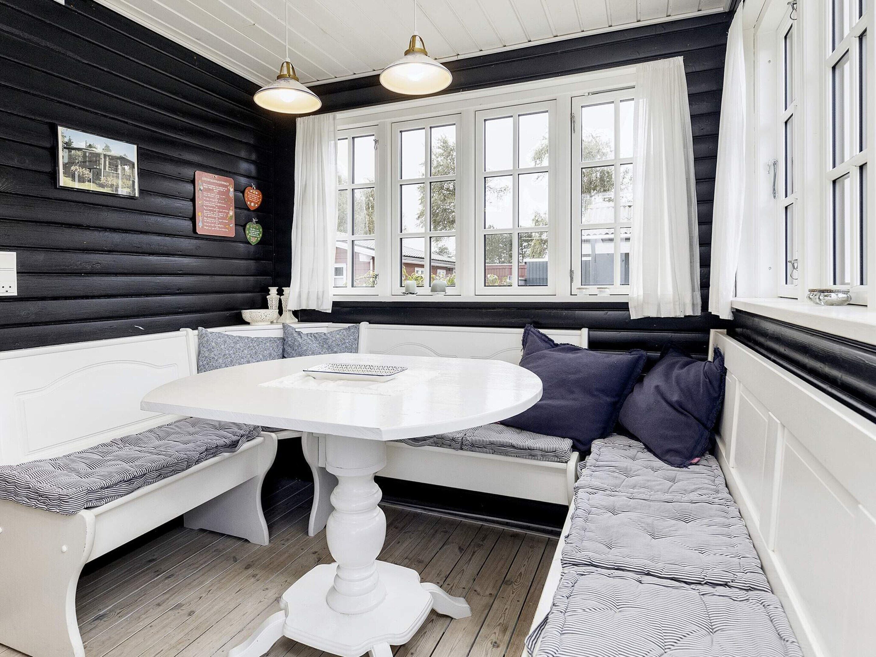 A room with a white round table and bench seating, a window with white curtains, and a black wall with a picture and a sign.