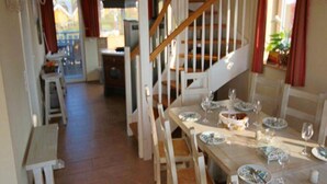 Ein Essbereich mit einem Holztisch, gedeckt für vier Personen, umgeben von weißen Stühlen. Eine Holztreppe mit Geländer führt zu einer erhöhten Ebene.