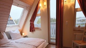 Ein Schlafzimmer mit schrägverdeckter Decke, einem Bett, einem Sessel und einem Fenster mit Blick auf ein Gebäude.