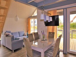 Una sala de estar moderna con un sofá, una mesa de comedor con sillas, un televisor de pantalla plana montado en la pared y ventanas amplias que ofrecen vistas al exterior.