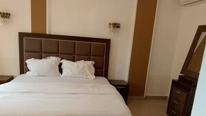 A neatly made bed with white linens, a dark headboard, and a wooden nightstand with a lamp. - Riyad City (Douala)